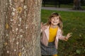 Toothless funny cute kid girl playing hide and seek in the autumn Park. Looks out from behind a tree with a playful smile and