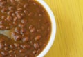 Top close view of a bowl filled with baked beans and onions in a white bowl with a spoon in the food atop a yellow tabletop