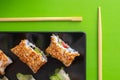 Top down view of a black square plate with multiple sushi rolls with sesame seeds and cucumber on green simple background with