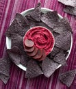 Top down view of a bowl of beetroot hummus surrounded by blue corn chips.