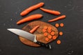 Top down view, chopping board, chef knife, and carrots, some of them cut in small circles, on black marble desk