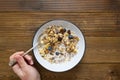 Top vie of hand holding spoon of oats for breakfast, granola with dried fruit and blueberry, milk and honey. Wooden background.