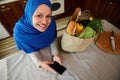 Top view Arab Muslim woman in hijab, smiles looking up at camera, checks expenses, using mobile phone with copy ad space