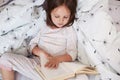 Top view of attentive pensive little kid holding book, reading with interest, lying in bed, being busy, doing her homework, Royalty Free Stock Photo