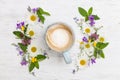 Top view on beautiful wild flowers  in shape of wreath and a cup of cappuccino coffee on white wooden background.  Summer flowers Royalty Free Stock Photo