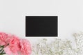 Top view of a black card mockup with gypsophila and a bouquet of pink carnations on a white table
