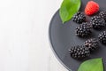 Top view of blackberries, one raspberry and green leaves on dark plate, on white wooden table, horizontal Royalty Free Stock Photo