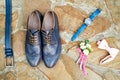 Top view of blue leather groom shoes, watches, belt, boutonniere, pink bowtie on brown natural stone. Groom wedding accessories.