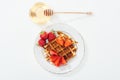 Top view of bowl with honey and wooden dipper near plate with waffle and strawberries on white.