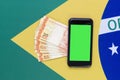 Top view of Brazilian green screen cell phone with Reail bills over Brazilian flag in the background. Copy space and green screen