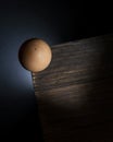 Top view of a chicken egg precariously balanced on the corner of a wooden table as it is about to fall to the ground