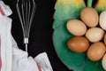 Top view of chicken eggs in a leaf shaped plate and an eggbeater on a black background