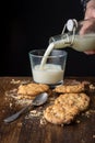 Top view of chocolate chip cookies, crumbs, spoon and hand with bottle pouring milk into glass cup, on wooden table and black back Royalty Free Stock Photo