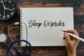 Top view of clock, stethoscope, and hand holding pen writing Sleep Disorder on notebook on wooden background