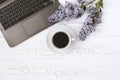 Top view of a computer and a cup of coffee and lilac flowers on a white wooden table. Place for text