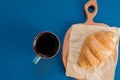 Top view of cup of black coffe and croissant on a cutting board and craft paper on blue background with copy space. Morning Royalty Free Stock Photo