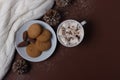 Top view of cup of cocoa with marshmallows, cookies, white sweater, pine cones and snow on brown background. Winter concept