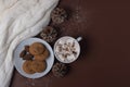 Top view of cup of cocoa with marshmallows, cookies, white sweater, pine cones and snow on brown background. Winter concept.