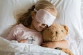 Top view of cute blonde little girl in pajamas and blindfold sleeping in white bed with teddy bear, awaking early in the morning
