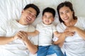 Top view of cute little boy and his asian parents looking at camera and smiling while lying on the white bed at home. Royalty Free Stock Photo