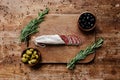 Top view of cutting board with rosemary, black and green olives in two bowls and delicious sliced salami on wooden table