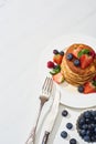 Top view of delicious pancakes with maple syrup, blueberries and strawberries on plate near fork and knife on marble white surface