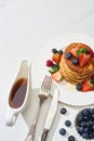 Top view of delicious pancakes with maple syrup, blueberries and strawberries on plate near fork and knife on marble white surface