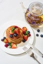 Top view of delicious pancakes with maple syrup, blueberries and strawberries on plate near herbal tea in teapot, fork and knife Royalty Free Stock Photo