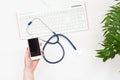 Top view doctor`s hand holding smartphone, stethoscope and keyboard on white table.