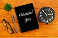 Top view of eyeglasses,pen,clock,plant and notebook written with Financial Year on wooden background.