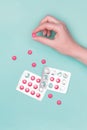 Top view of female hand holding pink prescription pill next to blister packs over pastel blue background.