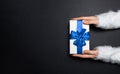 Top view of female hands giving a gift box with blue ribbon bow, wearing white sweater on textured background of black color. Royalty Free Stock Photo