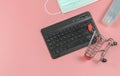 Flat lay of computer keyboard , shopping cart , alcohol sanitizer hand gel and medical face mask on pink background with copy