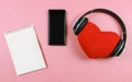 Flat lay of red heart pillow coverd with headphones, blank page opened note book and blank black screen mobile phone on pink Royalty Free Stock Photo