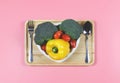 Flat lay of vegetables capsicum, broccoli and tomato in heart shape plate with fork and spoon in wooden tray on pink background