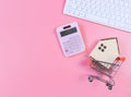 Flat layout of wooden house model in shopping trolley and pink calculator and computer keyboard  on pink  background with copy Royalty Free Stock Photo