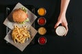 top view of french fries with delicious burger on tray, assorted sauces and hand holding glass of beer