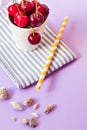 Top view of cherries on a stripped cloth and a yellow stripped straw with seashells on purple background