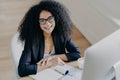 Top view of glad African American businesswoman smiles pleasantly, notes information, makes notes, wears transparent glasses,