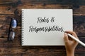 Top view of glasses and hand holding pen writing Roles and Responsibilities on notebook on wooden background