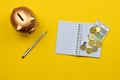 Top view of a golden piggy bank, coins, banknote, notebook, and pen on a yellow background