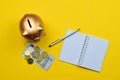 Top view of a golden piggy bank, coins, banknote, pen, and an open notebook on a yellow background