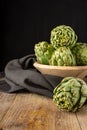 Top view of green artichokes in wooden bowl with black cloth, with selective focus, on wooden table, black background, vertical Royalty Free Stock Photo