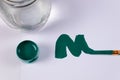 top view of a green smear in the form of the letter m and next to a green jar of gouache, as well as a transparent jar of water