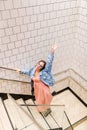 Top view of happy young woman on urban clothes climbing stairs. White tile wall. Fashion model wearing pink dress, denim jacket, Royalty Free Stock Photo