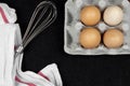 Top view of chicken eggs in a carton box and an eggbeater on a black background