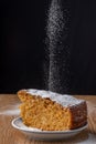 Top view of icing sugar falling on portion of carrot cake, in blue and white plate on wooden table with black background