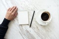 A hand going to write down on a blank white notebook with coffee cup on table