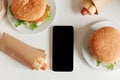 Top view image of junk food on table, tasty sausage in dough and big hamburgers, smart phone with black blank screen, copy space