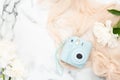 Top view instant film camera on marble background with white peony flowers and pastel color scarf. Minimal flat lay style
